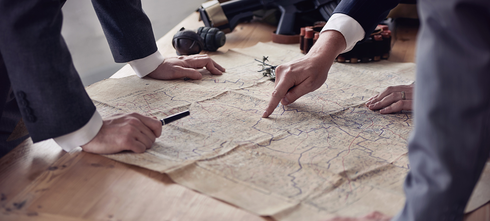 Three men in suits stand around a military map on a table, pointing out strategic targets (only their hands and arms are visible). Next to the map on the table, there is a hand grenade, a military-grade gun and bullets. Three men in suits stand around a military map on a table, pointing out strategic targets (only their hands and arms are visible). Next to the map on the table, there is a hand grenade, a military-grade gun and bullets.