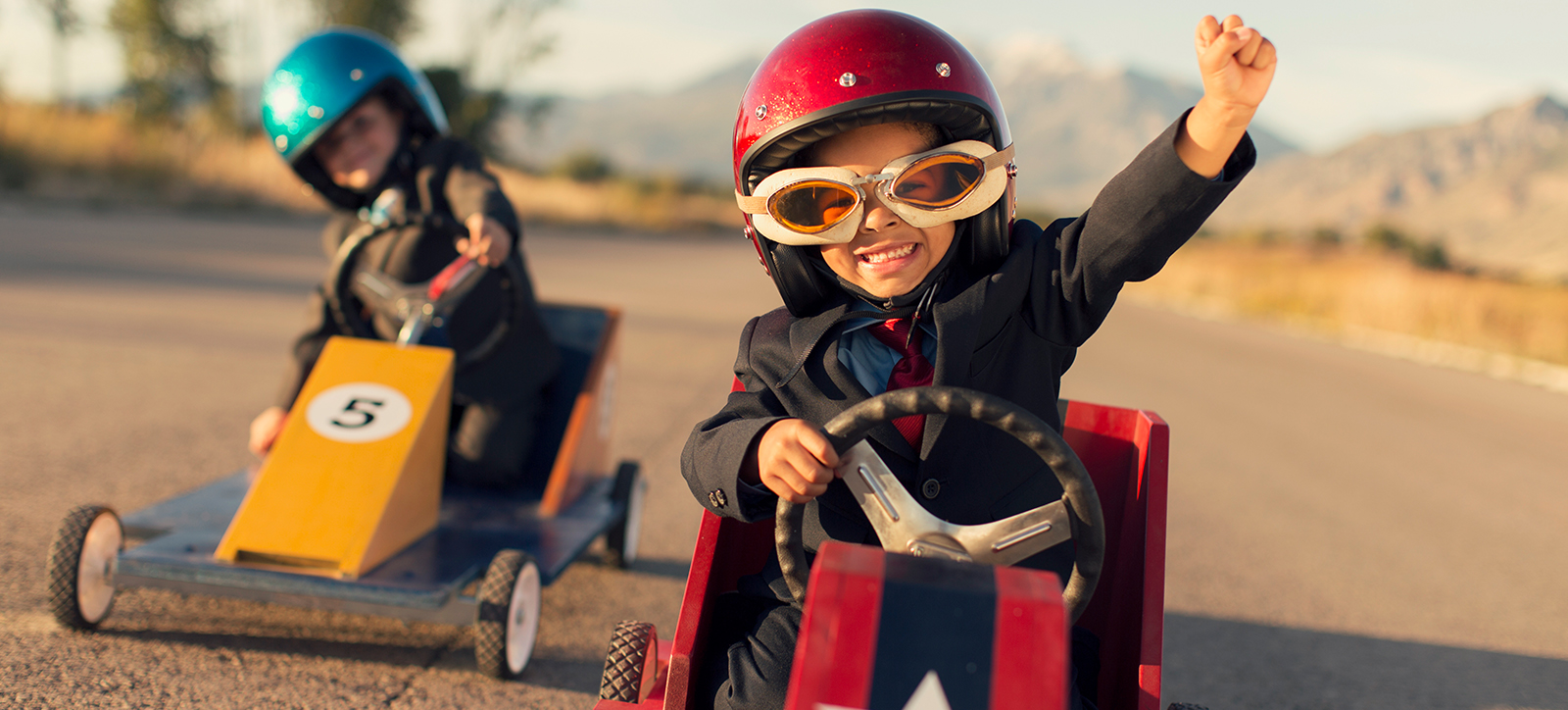 Two children in business suits drive go karts around a track, representing the race for competitive advantage between rival companies