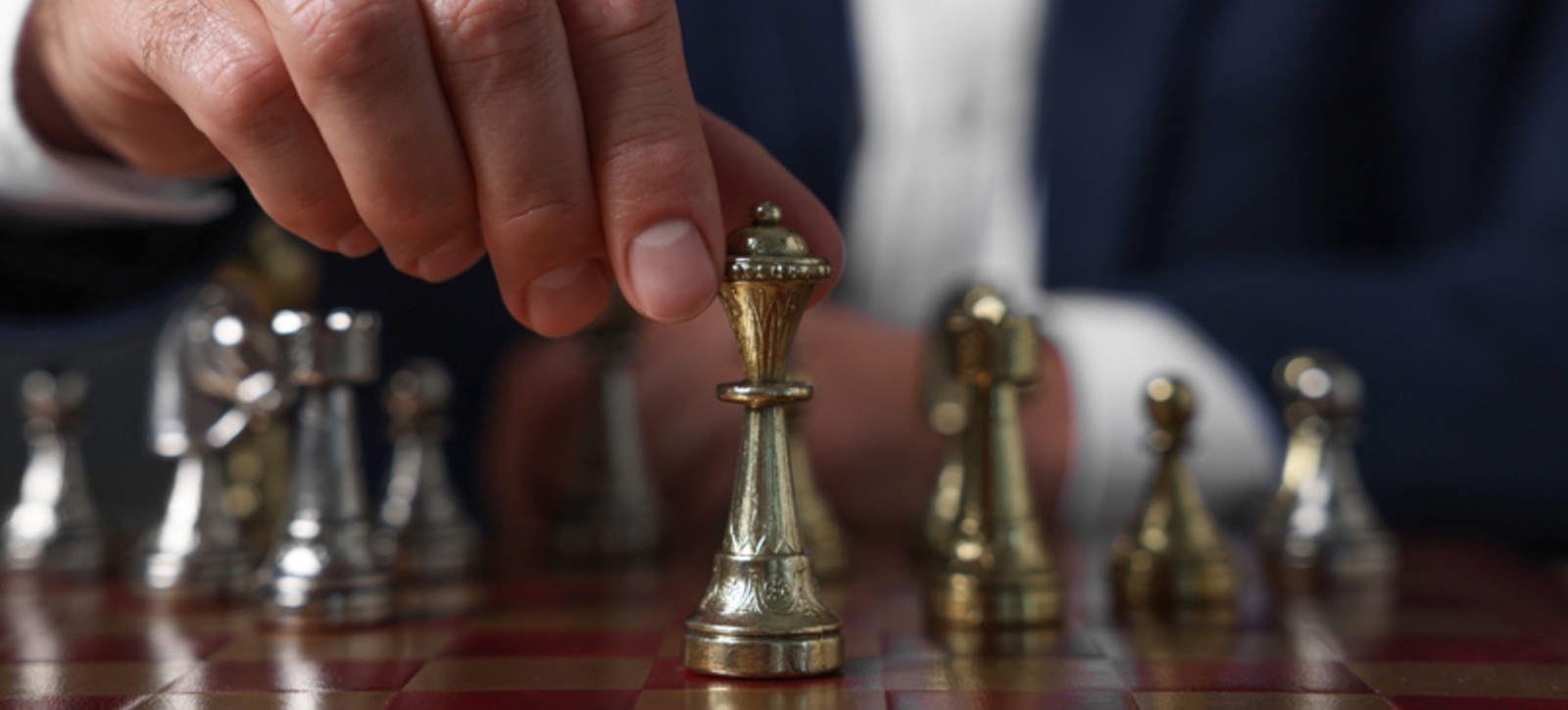 A close up photograph of a man in a business suit sat at a chess board, reaching out to move a piece.