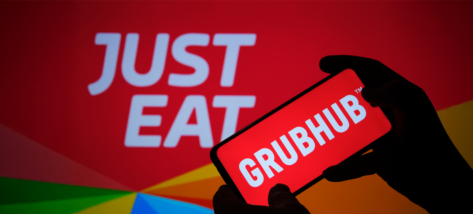 A silhouetted pair of hands hold a mobile phone displaying the name of US takeaway delivery service Grubhub. In the background is a giant banner showing the Just Eat logo.