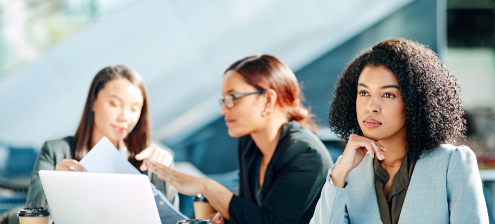 A Black office worker sits on the periphery of a meeting, starting into space, as she is ignored by her colleagues.