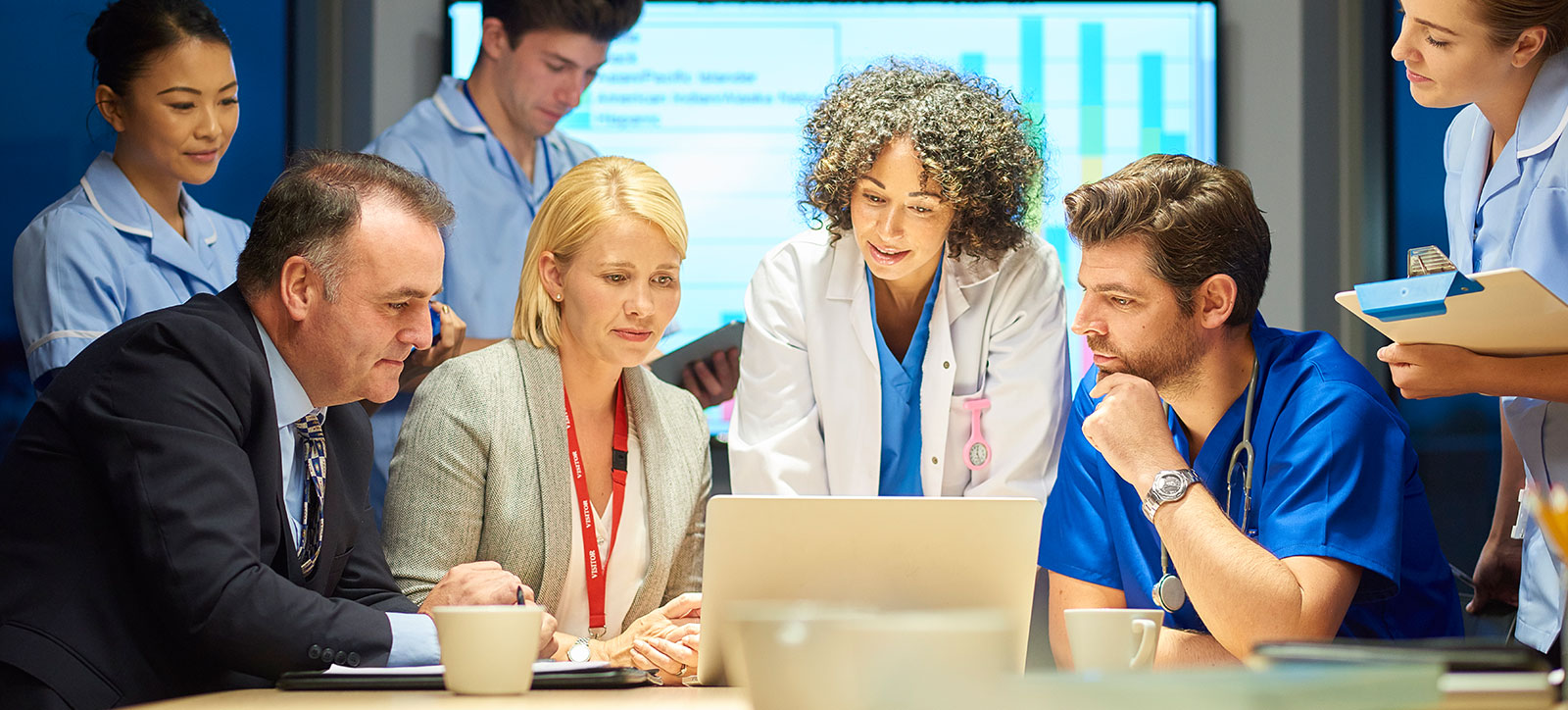 A team of doctors, nurses, and NHS managers sit around a computer in a board room as they assess the social and economic value of a continuous improvement initiative.