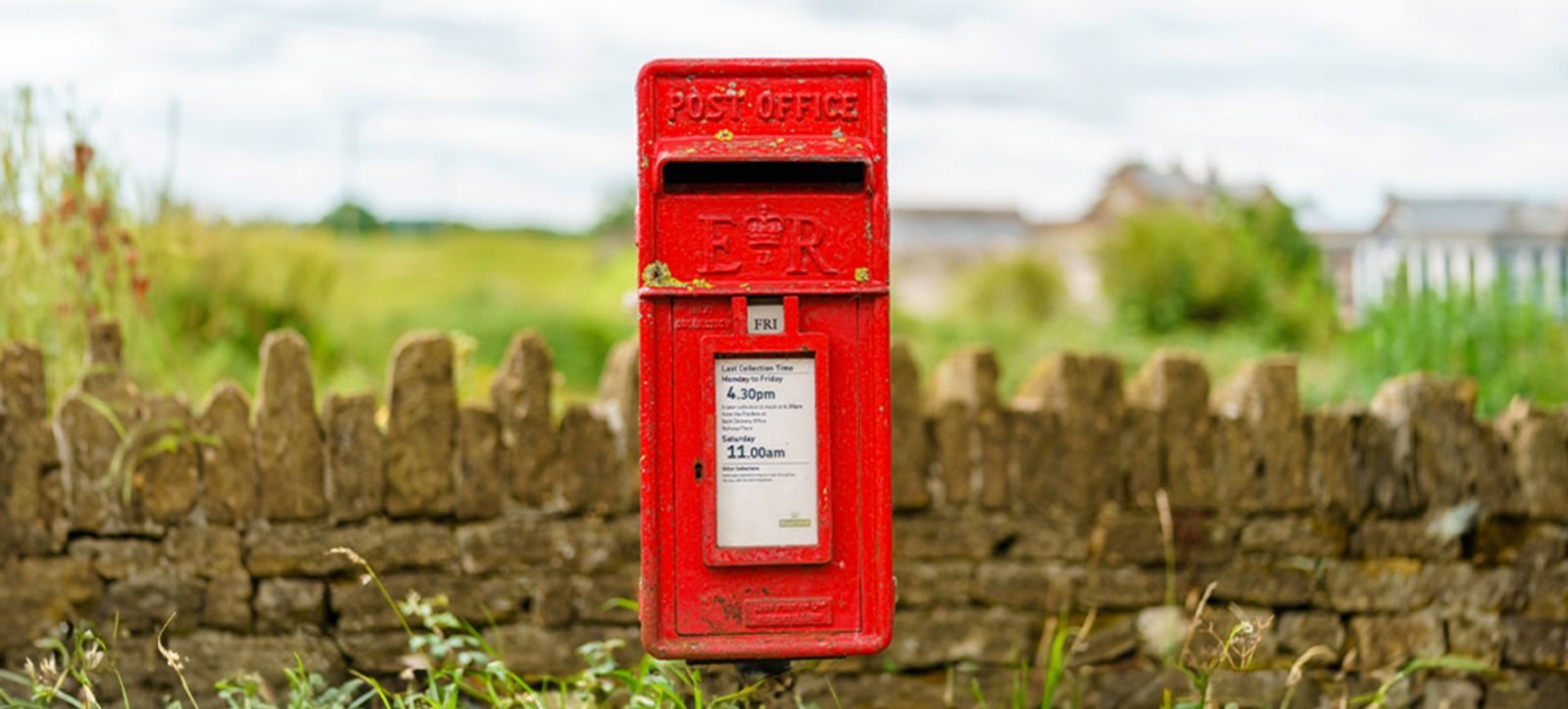 A photograph of a weathered Royal Mail post box in front of a rural stone wall.