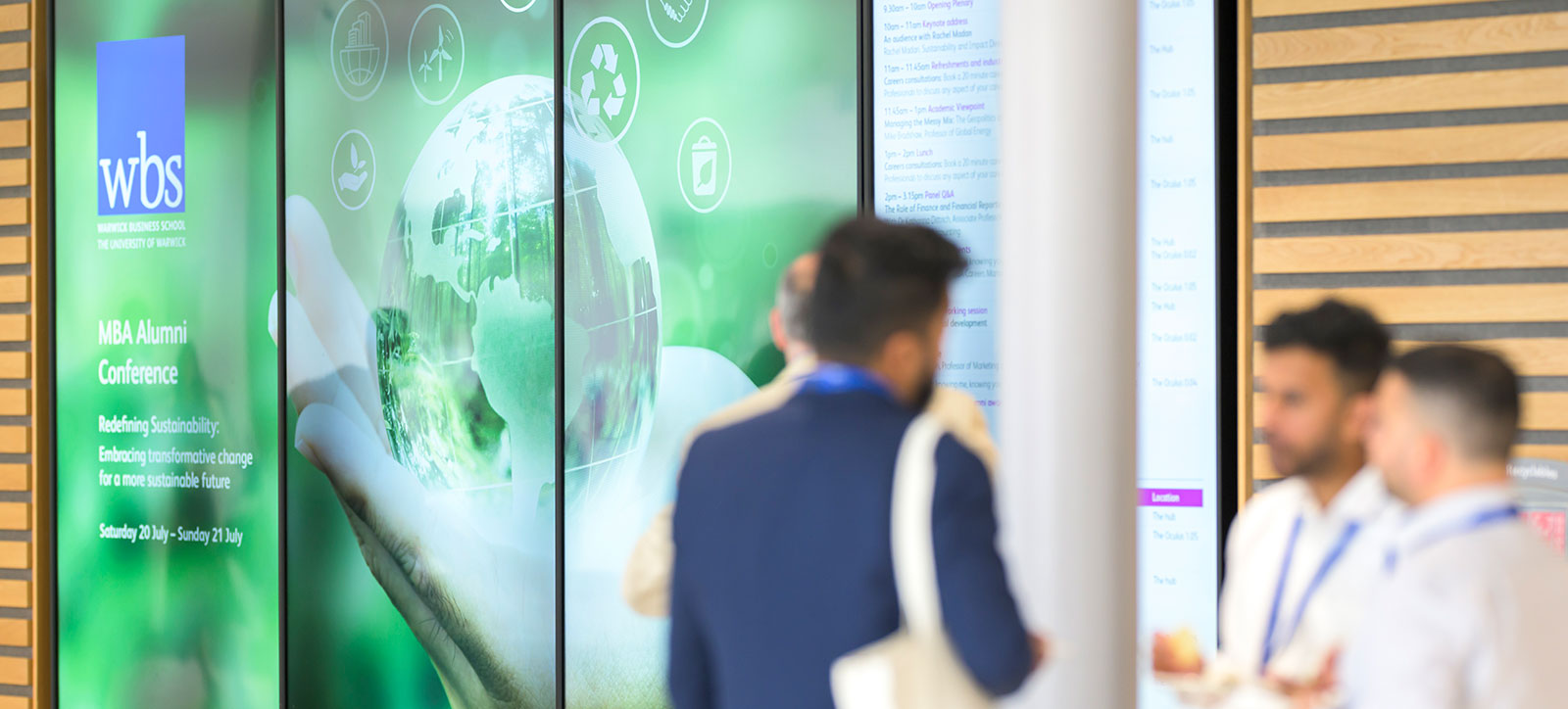 A group of Warwick Business School alumni network in front of the giant videos screens at the annual MBA Alumni Conference. The screens show an image of a hand holding a hologram projected image of the Earth to illlustrate the conference's theme of sustainability.