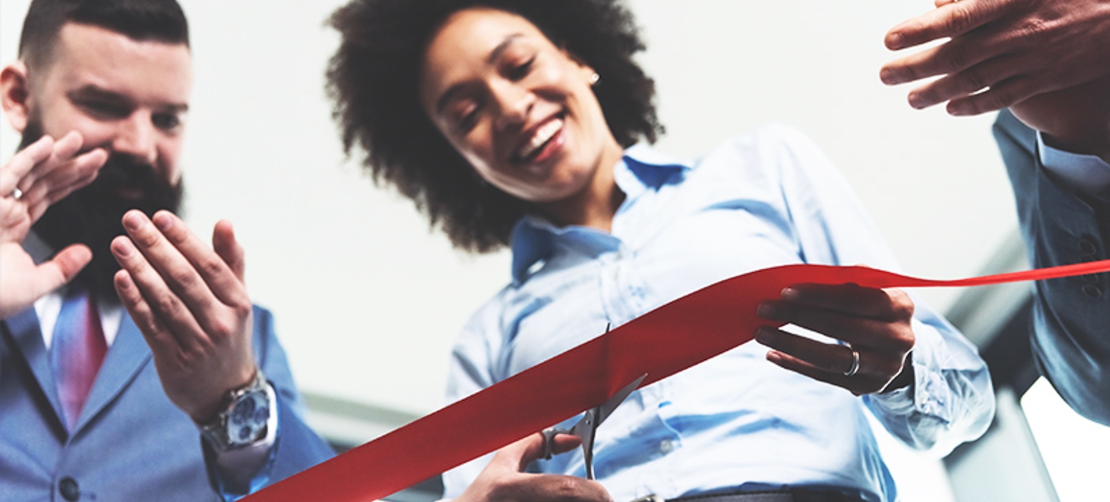 A young female entrepreneur, smartly dressing in a light blue shirt and grey skirt, cuts a bright red ribbon to open a new business. A young businessman in a suit, with short hair and a bear, stands next to her applauding. A young female entrepreneur, smartly dressing in a light blue shirt and grey skirt, cuts a bright red ribbon to open a new business. A young businessman in a suit, with short hair and a bear, stands next to her applauding.