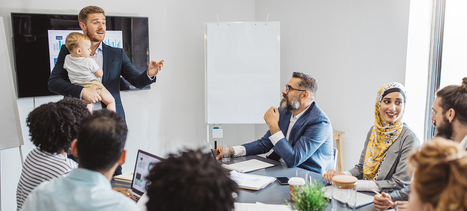 A male manager leads a boardroom meeting while holding a baby.