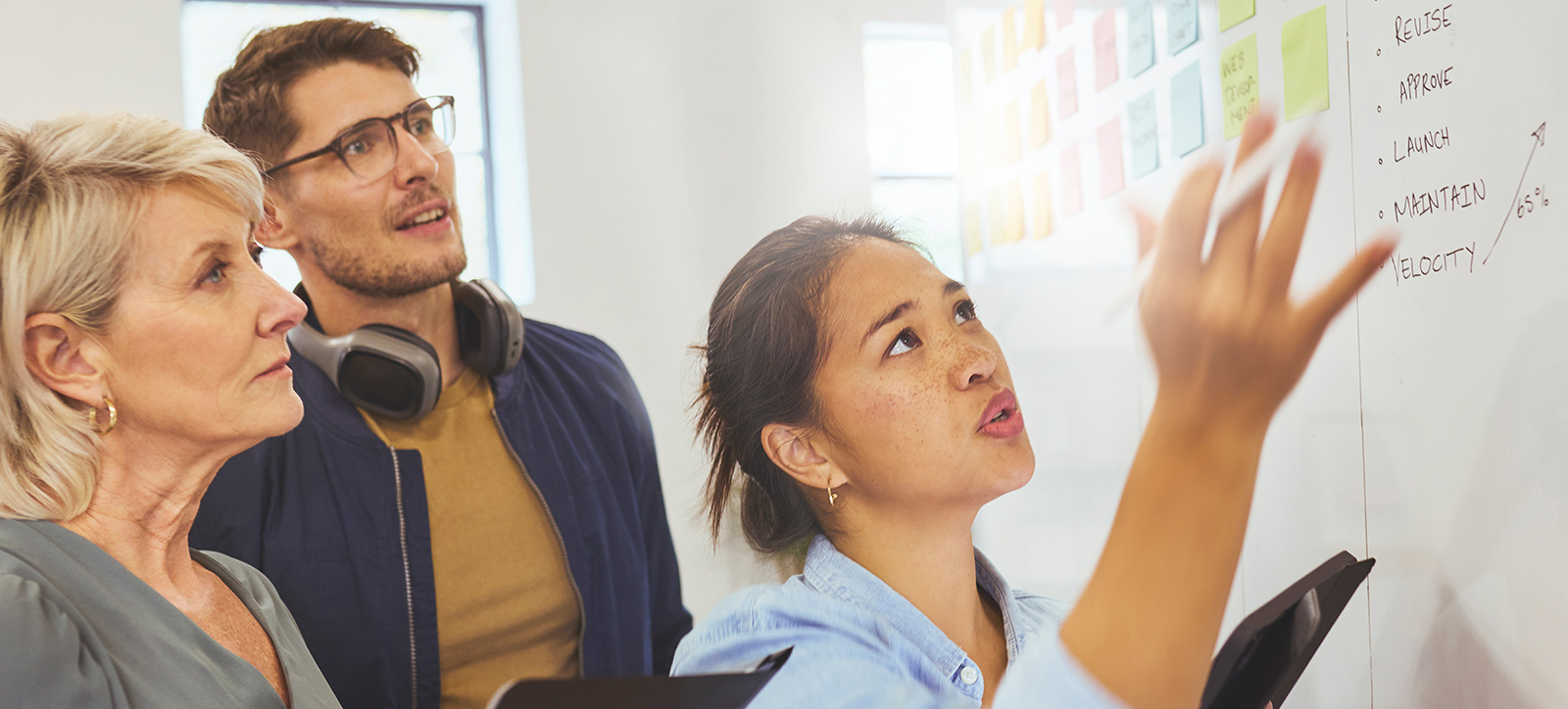 A young woman points to a whiteboard with an older lady and man looking at it