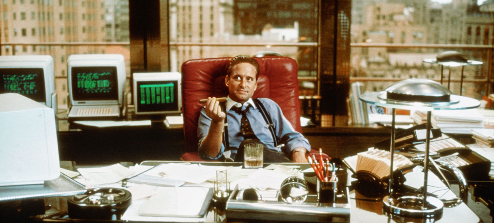 An image of Michael Douglas in his Oscar-winning role as Gordon Gekko in Wall Street. He is sat in a padded, red leather chair and is holding a cigar. A glass of whiskey rests on the desk in front of him. The New York skyline is visible through the window behind him. An image of Michael Douglas in his Oscar-winning role as Gordon Gekko in Wall Street. He is sat in a padded, red leather chair and is holding a cigar. A glass of whiskey rests on the desk in front of him. The New York skyline is visible through the window behind him.
