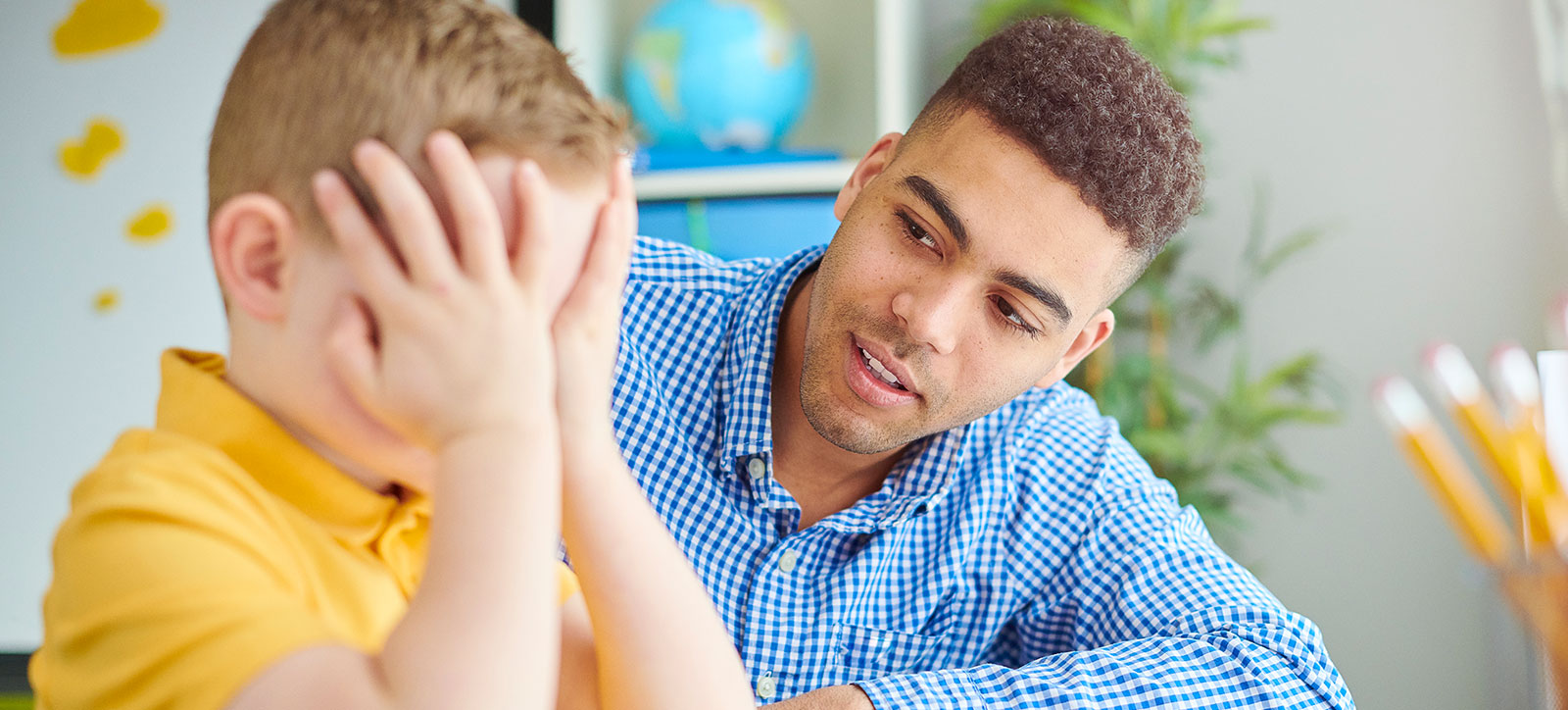 A young male teacher reassures a primary school boy who is sat with his head in his hands, looking distressed. A young male teacher reassures a primary school boy who is sat with his head in his hands, looking distressed.