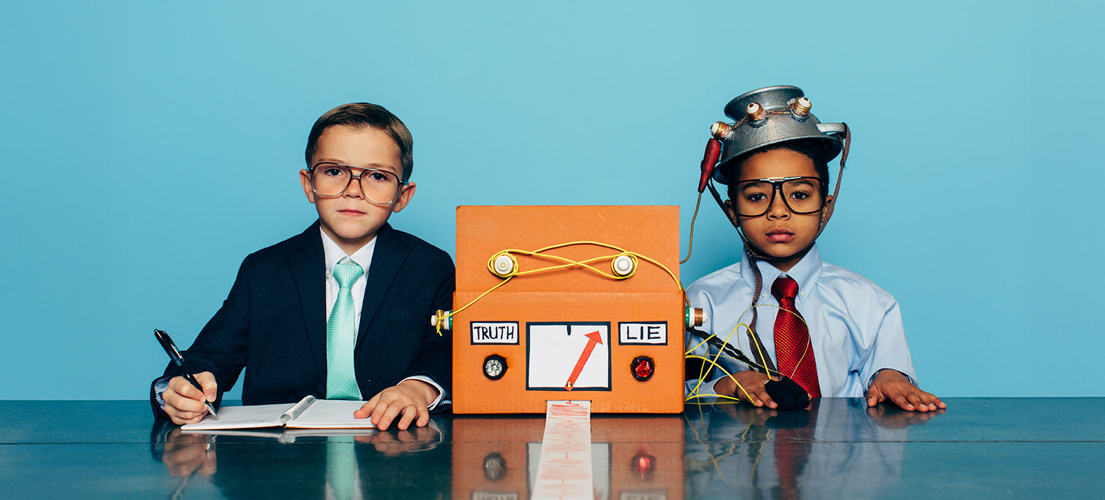 Two boys sat side by side at a desk. Both are dressed as office workers, wearing shirts and ties. One is fitted to a homemade, pretend lie detector. He has an upturned colander on his head, with wires connected it to a decorated cardboard box. A long strip of paper with a red polygraph line drawn on is emerging from the box. The other boy is wearing a suit jacket to make him look more like an authority figure and has a pen in his hand and is recording the results in a notebook. Two boys sat side by side at a desk. Both are dressed as office workers, wearing shirts and ties. One is fitted to a homemade, pretend lie detector. He has an upturned colander on his head, with wires connected it to a decorated cardboard box. A long strip of paper with a red polygraph line drawn on is emerging from the box. The other boy is wearing a suit jacket to make him look more like an authority figure and has a pen in his hand and is recording the results in a notebook.