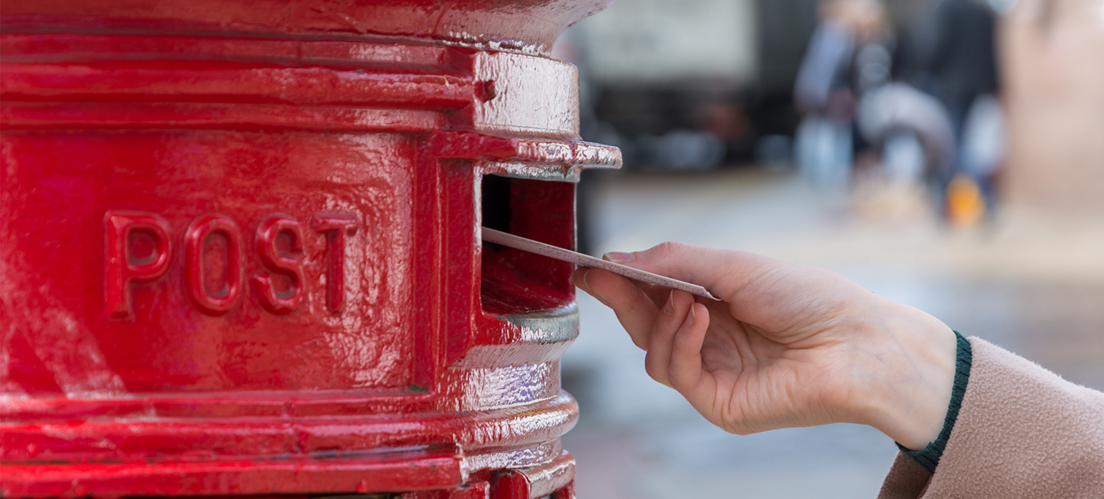 A close up of a woman's hand post a small stack of letters into a bright red Royal Mail postbox. The woman is wearing a dark, woolen sweater and a beige coat. The high street in the background is in soft focus, making it impossible to see individual details. A close up of a woman's hand post a small stack of letters into a bright red Royal Mail postbox. The woman is wearing a dark, woolen sweater and a beige coat. The high street in the background is in soft focus, making it impossible to see individual details.