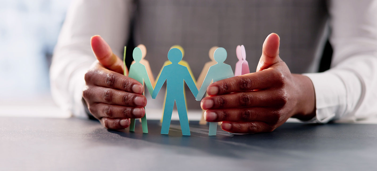 A close up of a leader's hands protectively surrounding a circle of different coloured paper figures of people, representing how organisations can foster inclusive leadership.